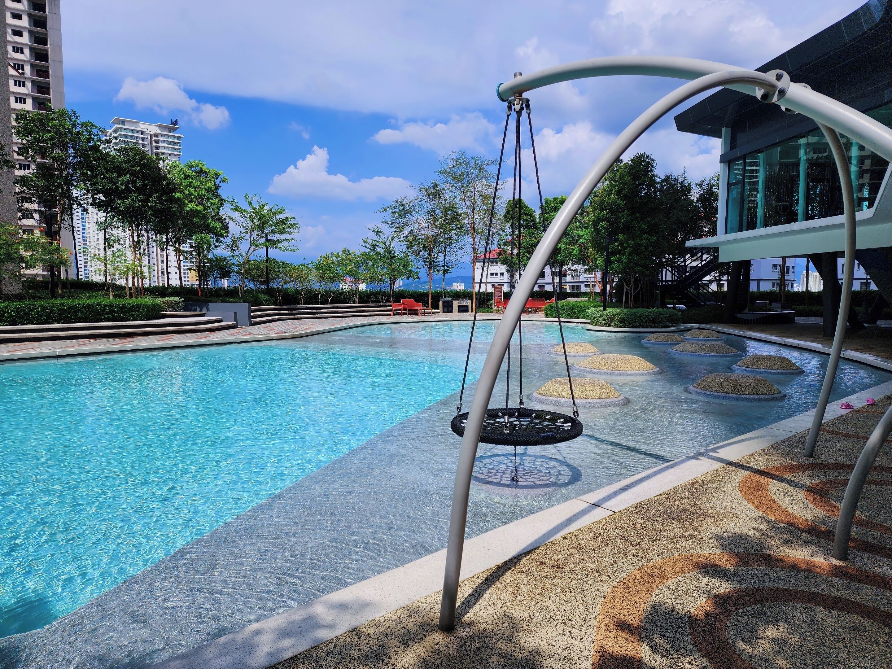 A swimming pool with a swing chair, surrounded by a landscaped area with trees and a view of buildings in the background.