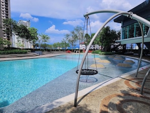 A swimming pool with a swing chair, surrounded by a landscaped area with trees and a view of buildings in the background.