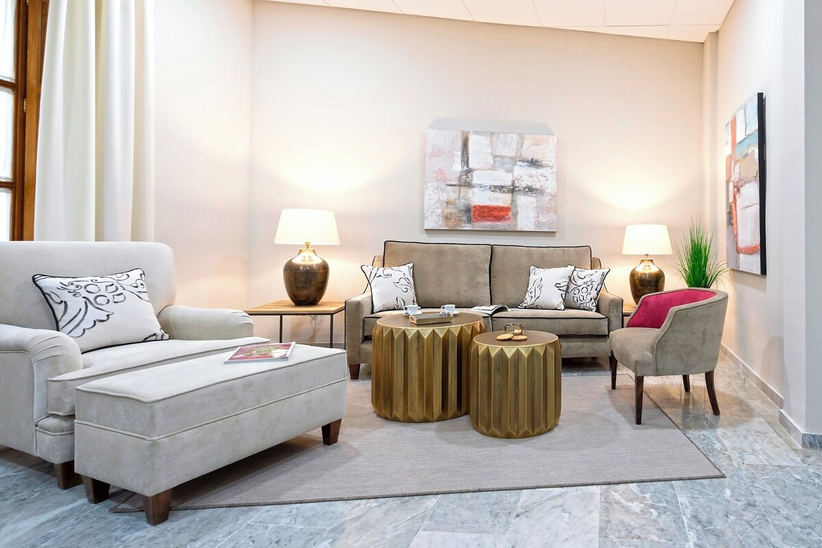 Una sala de estar moderna con un sofá, un sillón, un reposapiés, dos mesitas de noche con lámparas, y un cuadro abstracto grande en la pared.