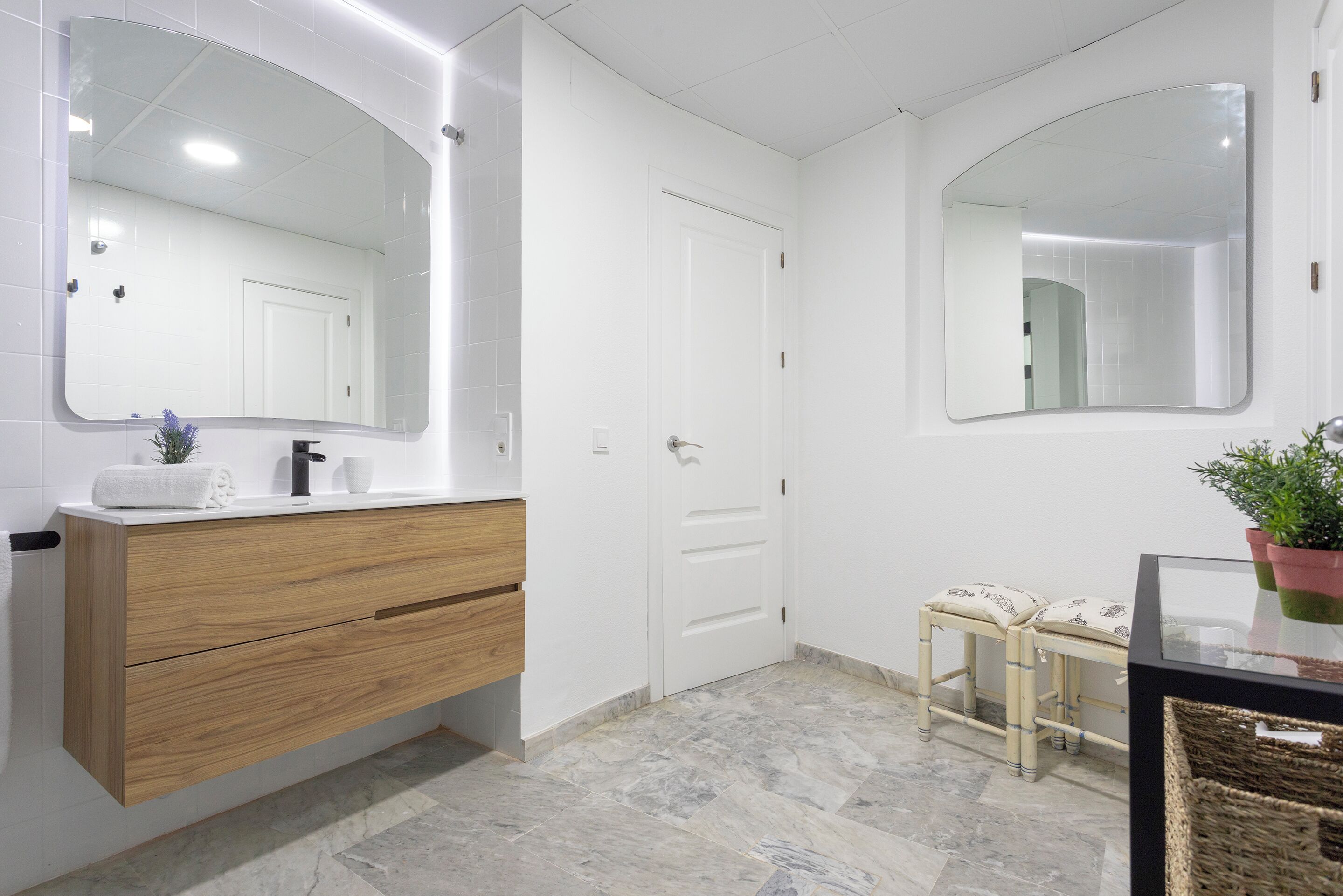 A modern bathroom with a wooden vanity, two mirrors, a white door, and a small bench.