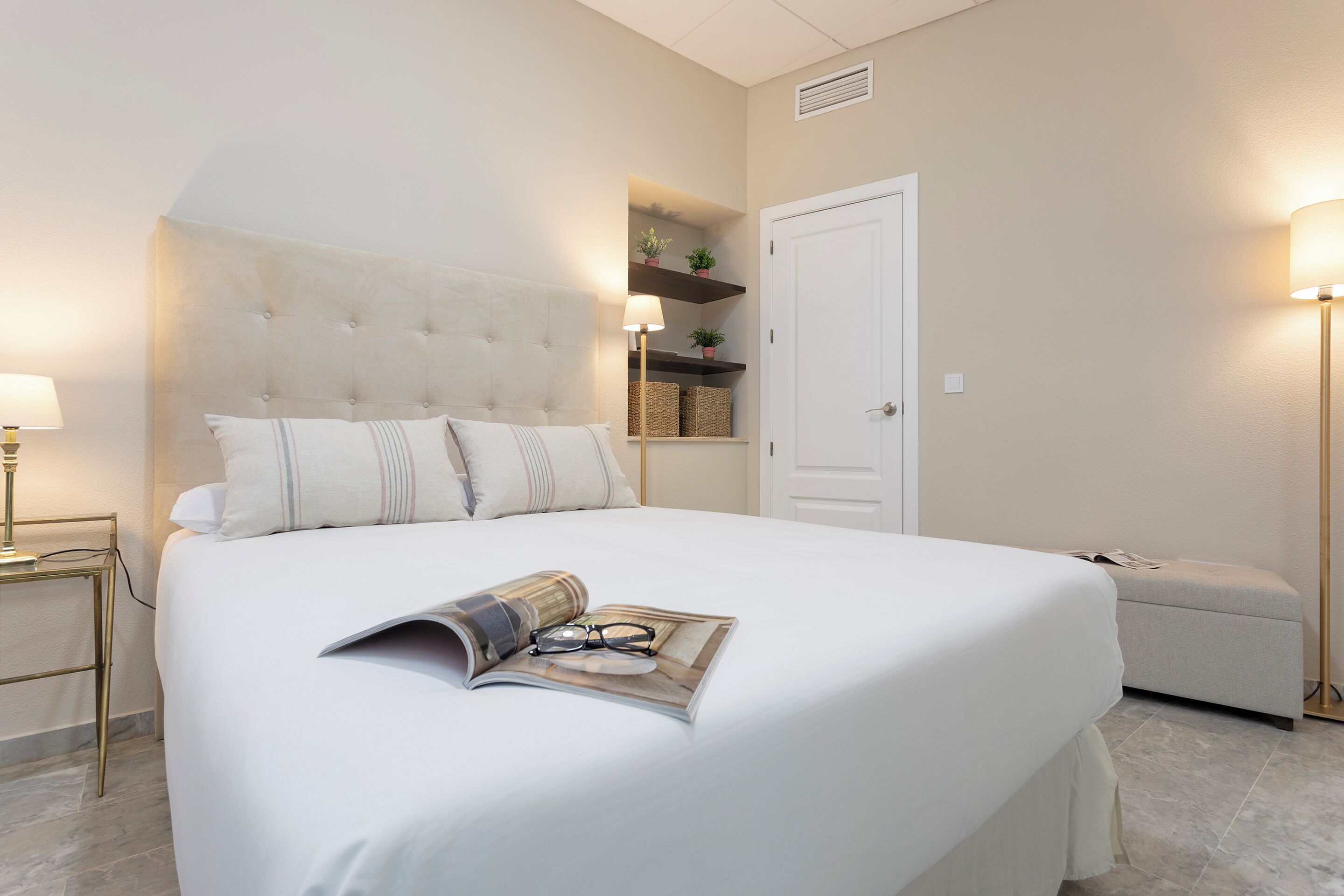 A neatly made bed with a magazine and glasses on it, a bedside lamp, a small table with a lamp, and a bench.