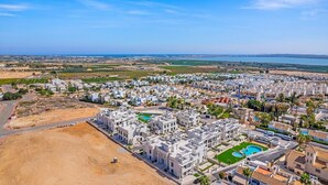An aerial view of a coastal residential area with modern buildings, a large swimming pool, and a clear view of the sea.