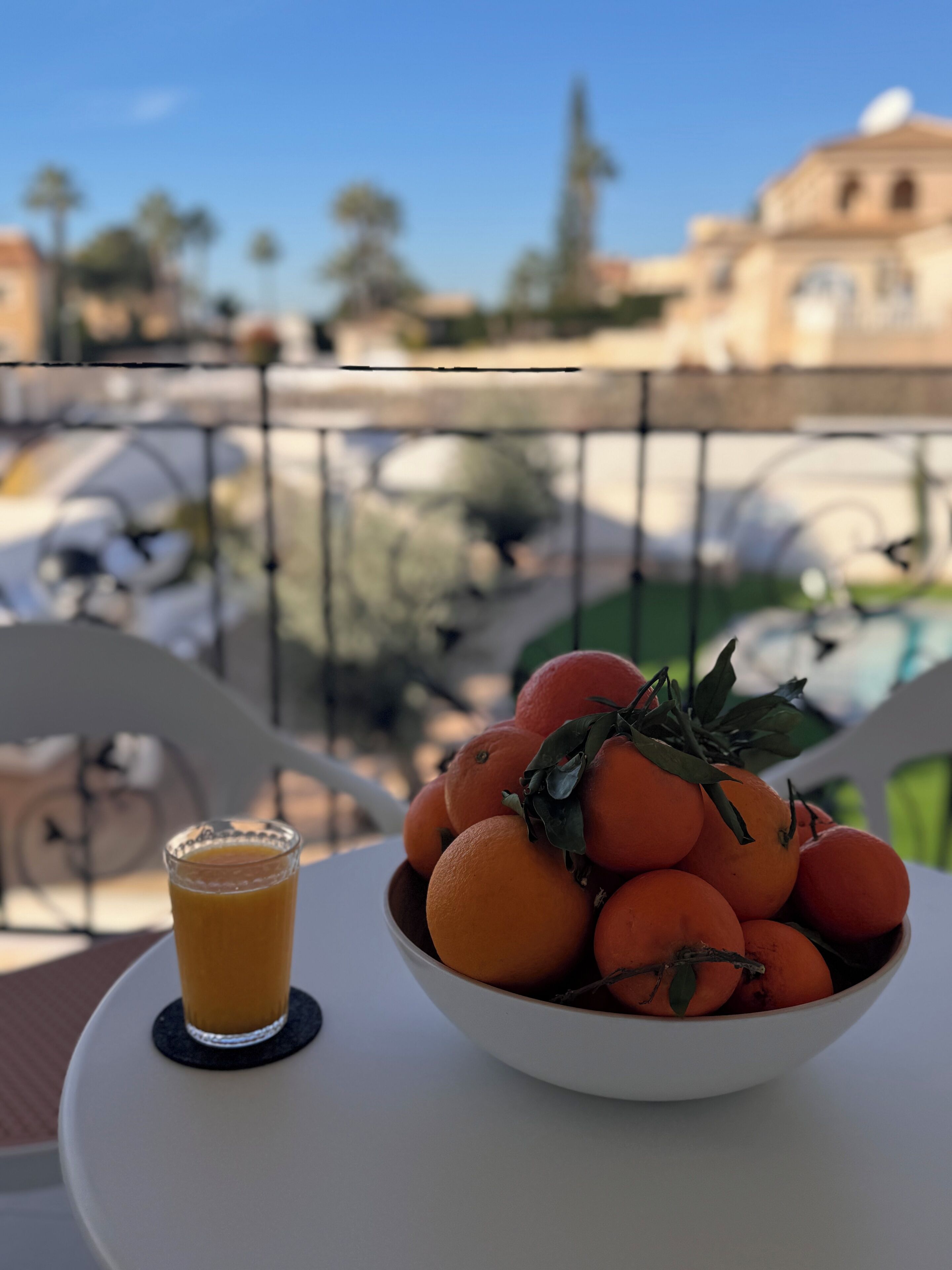 A bowl of oranges on a table with a glass of orange juice beside it, set on a balcony with a railing and buildings in the background.