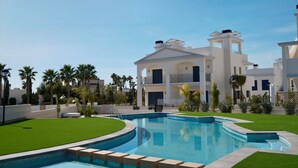 A modern hotel with a large swimming pool, surrounded by palm trees and a well-maintained lawn.