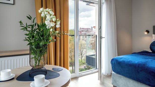 Une chambre d’hôtel avec un lit, une table avec des tasses et un vase de fleurs, et une vue sur les bâtiments à travers une grande fenêtre.