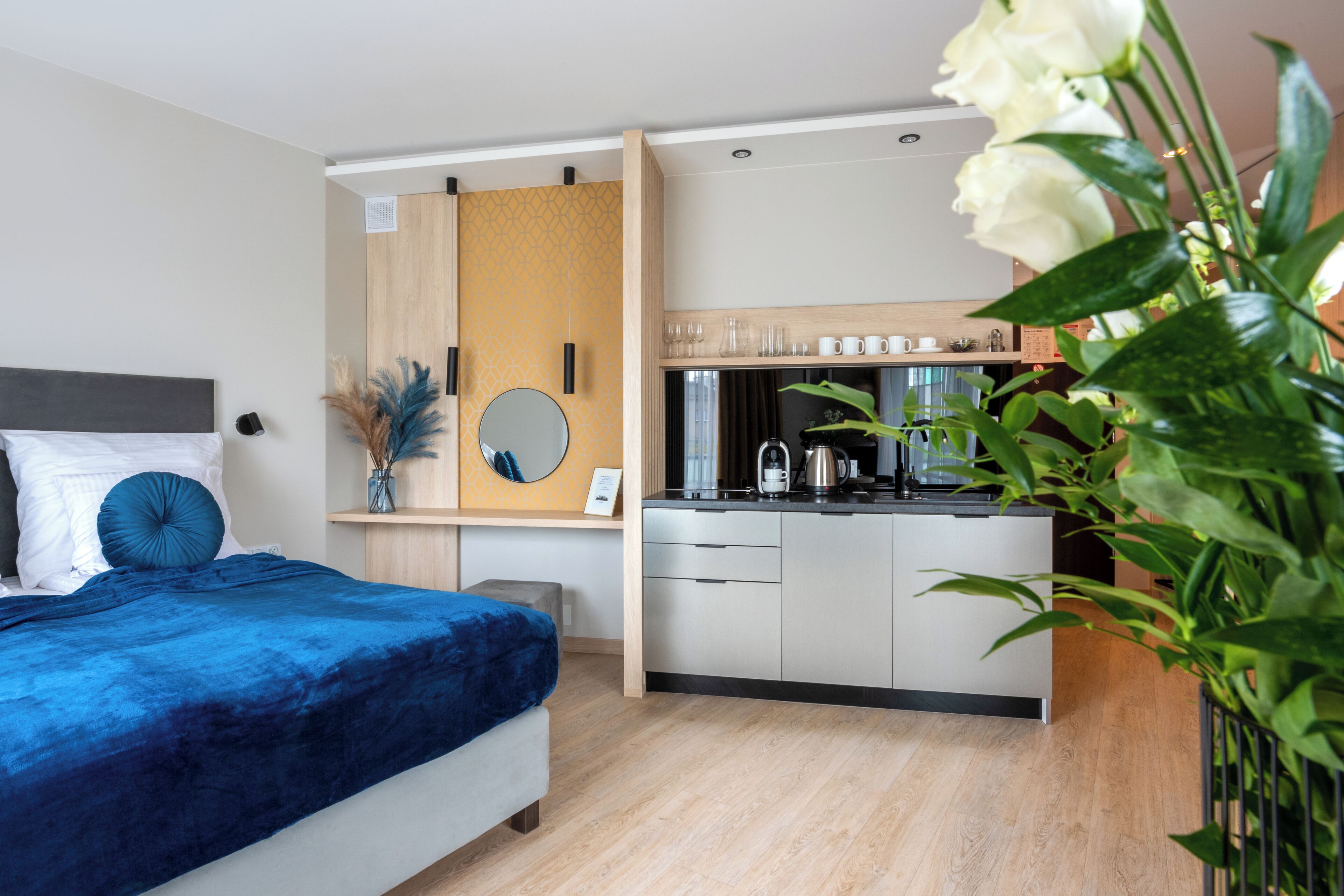 A modern hotel room with a bed, bedside table, mirror, and a kitchenette area.