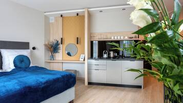 A modern hotel room with a bed, bedside table, mirror, and a kitchenette area.