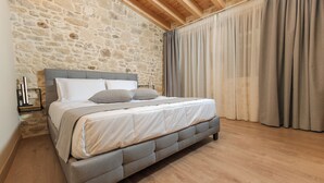 Een slaapkamer met een stenen muur, een bed met grijze beddengoed, houten vloeren en een groot raam met gordijnen.