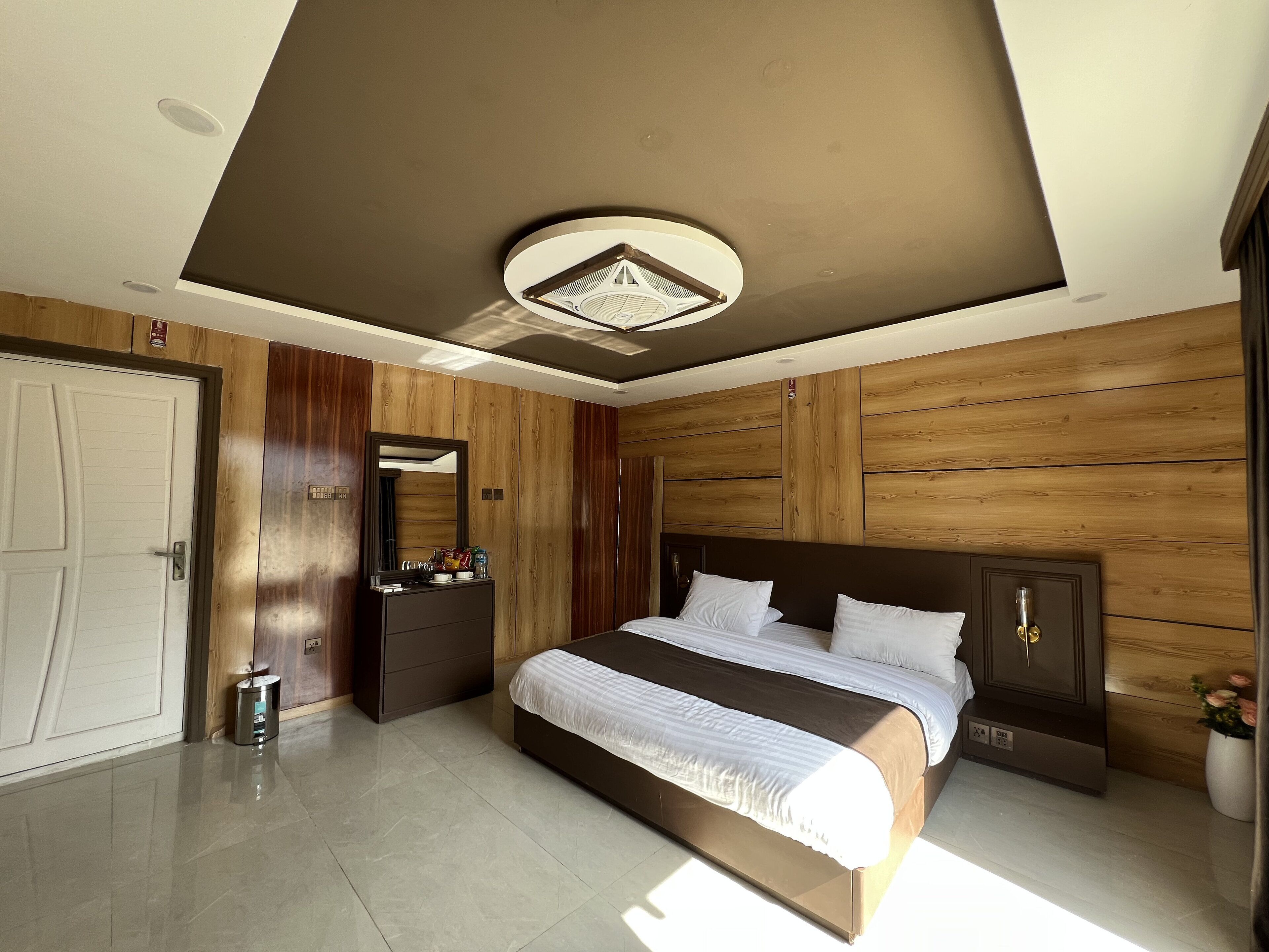 A modern bedroom with a large bed, wooden panel walls, a ceiling light fixture, and a small trash bin.