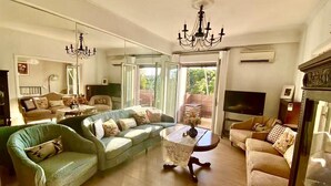 Un salón amplio con un sofá modular grande, una mesita de centro redonda y un televisor. Un candelabro cuelga del techo, y la habitación tiene ventanas grandes que dejan entrar luz natural.