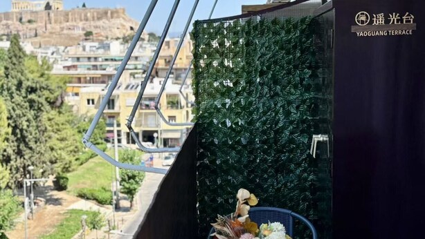 Un balcón con vistas a un paisaje urbano, una pared verde y frondosa, y una mesa con un arreglo floral.
