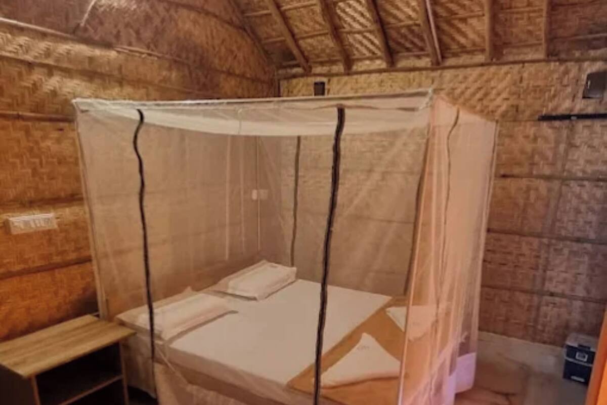 Un lit simple avec une moustiquaire, dans une pièce aux murs en bois et au toit de chaume.