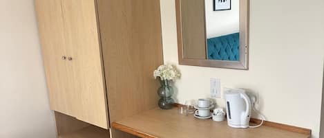 A compact room with a wooden desk, a mirror, a small vase with flowers, a kettle, and a bed.