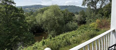 Un balcon donnant sur une rivière, entouré d’une végétation luxuriante et avec des collines visibles au loin.