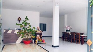 Un moderno vestíbulo de hotel con una recepción, una gran planta en maceta, una zona de comedor con sillas de madera y un altar decorativo.