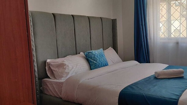 Ein ordentlich bezogenes Bett mit einer blau-weißen Tagesdecke, einem grau gepolsterten Kopfteil und einem Fenster mit blauen Vorhängen.