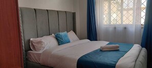 A neatly made bed with a blue and white bedspread, a grey upholstered headboard, and a window with blue curtains. - Perfect Palm Condos- N12 (Nakuru)