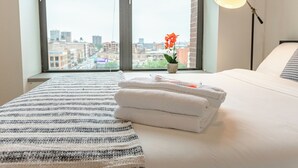 A hotel room with a large window, a bed with white linens, a striped area rug, a small potted plant with an orange flower, and a floor lamp. - McCormick South Loop 2BR Suite Near Soldier Field & Museum Campus (Chicago)