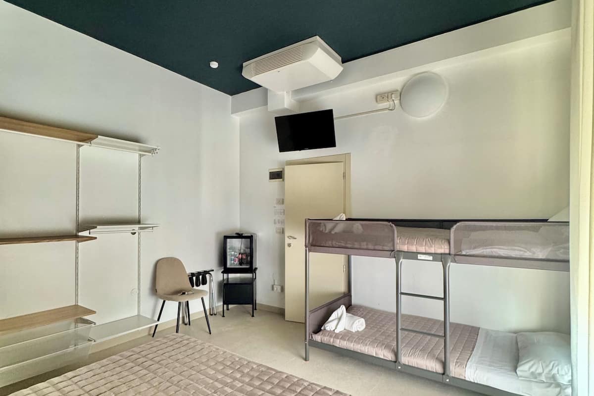 Habitación de hotel con una cama grande, una cama literera, un escritorio con silla, televisión y aire acondicionado instalado en el techo.