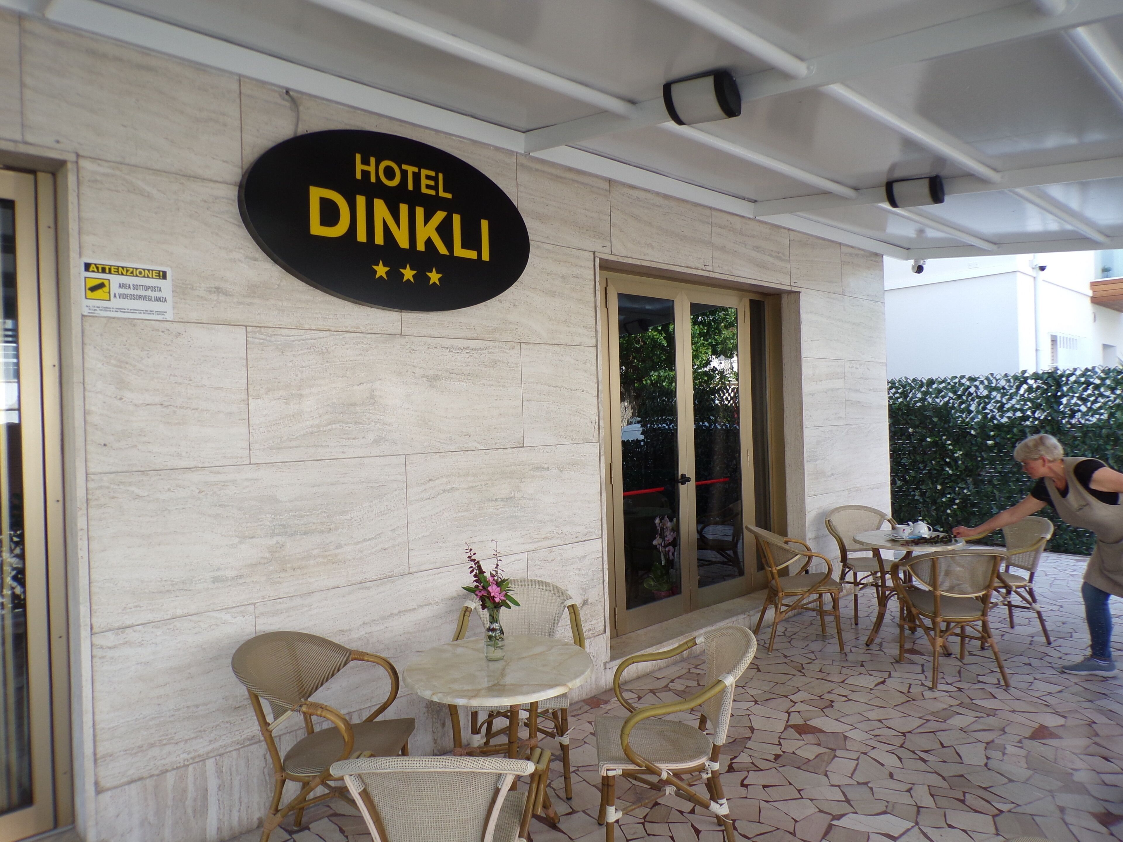 Ein Hotel mit Fassade, auf dem ein Schild mit dem Schriftzug 'HOTEL DINKLI' zu sehen ist. Draußen stehen Tische und Stühle, und eine Person kümmert sich um das Areal.