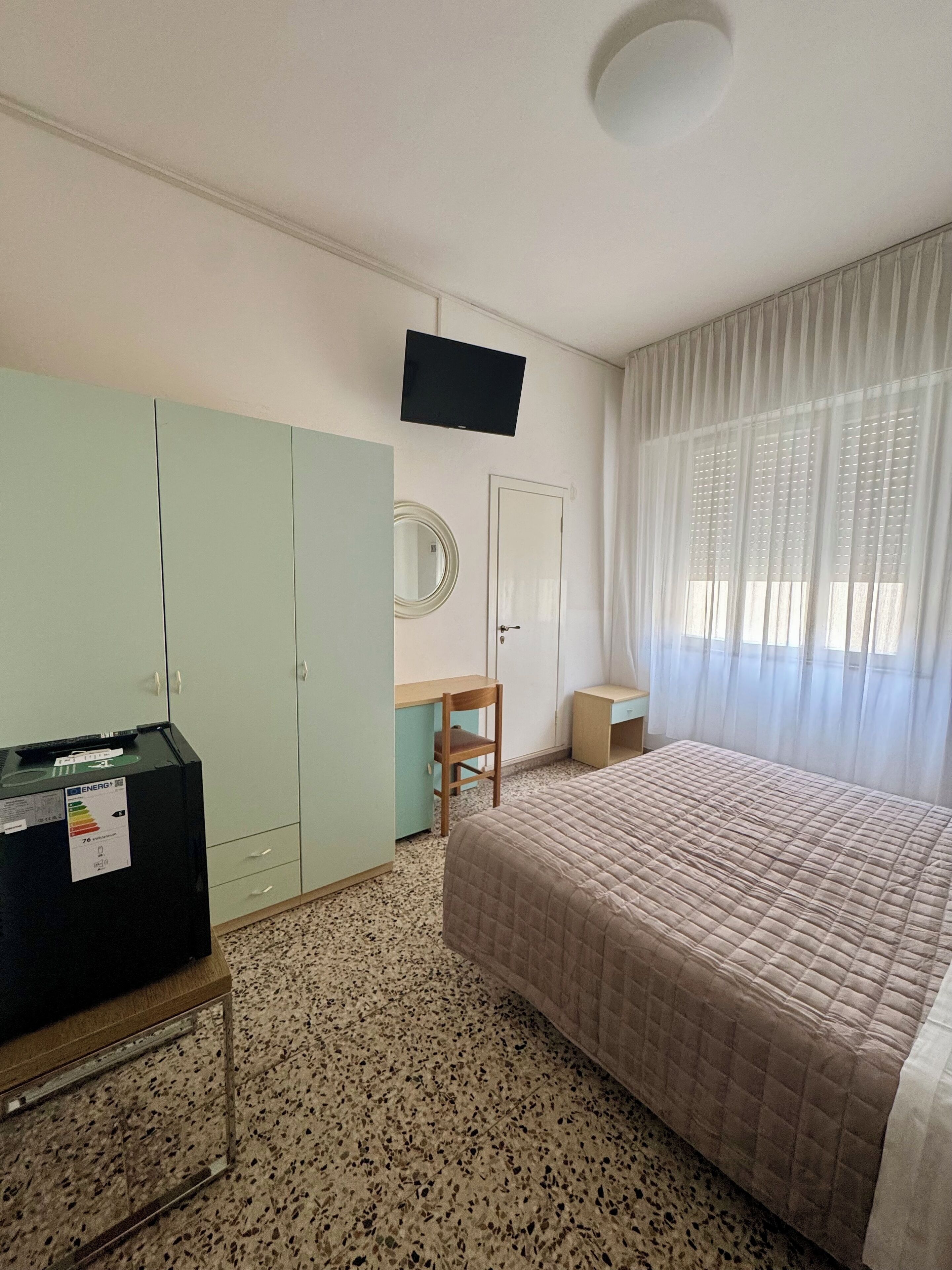 A hotel room with a bed, a desk, a wardrobe, a television, and a window with curtains.