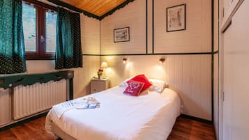 Ein ordentlich bezogenes Bett mit weißen Leinentüchern und einem roten Kissen, ein kleiner Nachttisch mit einer Lampe und ein Fenster mit Vorhängen.