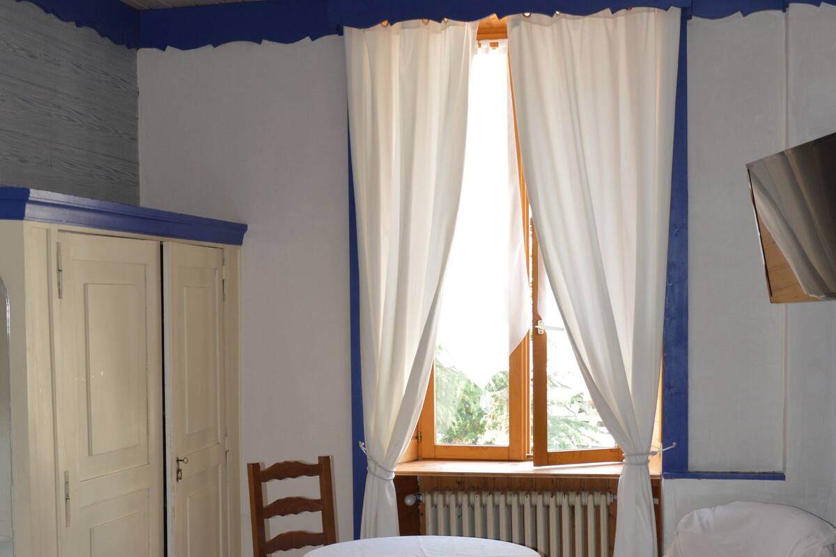 Une chambre avec deux lits, une table ronde et une chaise. On y trouve une fenêtre avec des rideaux, un radiateur et une armoire.