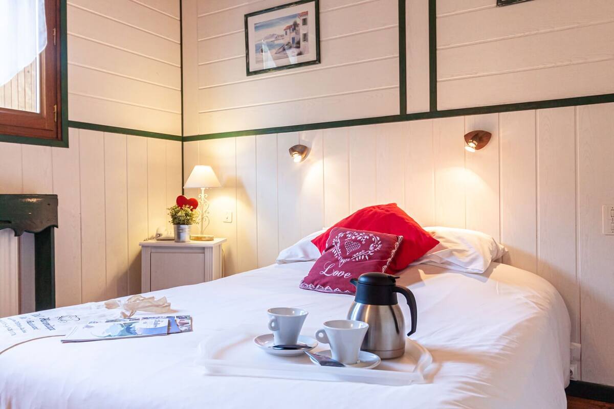 Un lit bien fait, avec un plateau contenant du café et des tasses, un oreiller rouge et une petite table sur laquelle se trouvent une lampe et des fleurs.