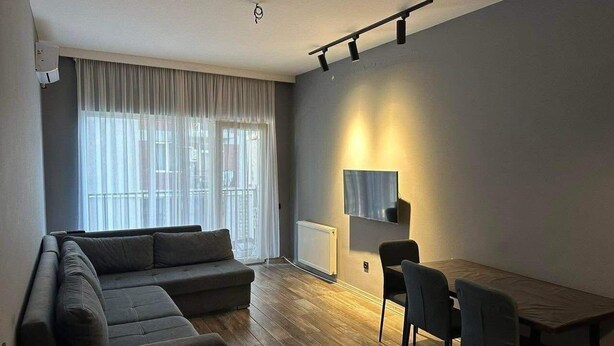 Ein modernes Wohnzimmer mit Ecksofa, Esstisch mit Stühlen, einem Wand-Fernseher und einem großen Fenster mit leichtem Vorhang.