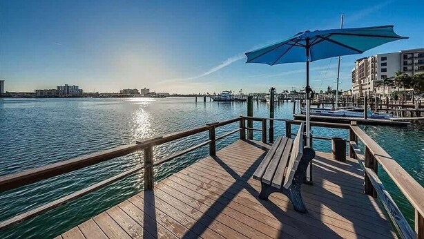 Eine Holzstege mit einer Bank und einem blauen Sonnenschirm, mit Blick auf ruhiges Wasser, im Hintergrund sind Gebäude zu sehen.