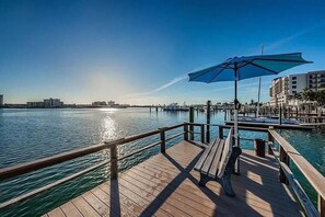 A wooden pier with a bench and a blue umbrella, overlooking a calm body of water with buildings in the background.