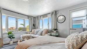 A modern bedroom with a large bed, a smaller sofa, a bedside table, and a view of the city through large windows. - Right on Clearwater Beach! Corner Suite Waterview (Clearwater)