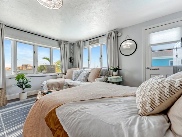 A modern hotel room with a large bed, a sofa, a small table, and a view of the city through large windows. - Water-view studio, heated pool, walk to beach (Clearwater)