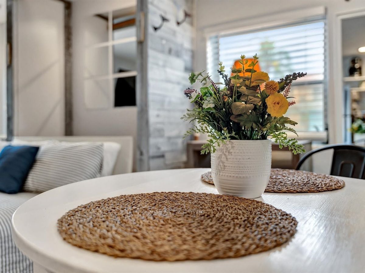 A dining table with woven placemats, a white vase with flowers, and a modern interior with a brick wall and a window.