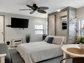 Ein Schlafzimmer mit Bett, Schreibtisch, Stuhl, Deckenventilator, einem an der Wand montierten Fernseher und einem Fenster mit Jalousien.