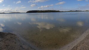 A calm lake with a sandy shore, a distant forested island, and a clear sky. - 8 Person Holiday Home in Maribo (Maribo)