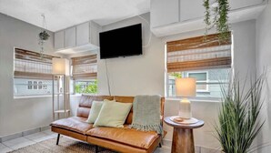 A modern living room with a leather sofa, a flat-screen TV mounted on the wall, and large windows with blinds. - Resort Condo just 2 Blocks from Clearwater Beach! (Clearwater)