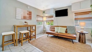 A living room with a sofa, wooden table, and chairs. A TV is mounted on the wall, and there is a window with blinds. - Resort Condo just 2 Blocks from Clearwater Beach! (Clearwater)