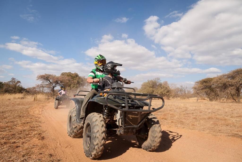 Una persona guida un ATV su un sentiero sterrato, con vegetazione rada e alberi sullo sfondo.