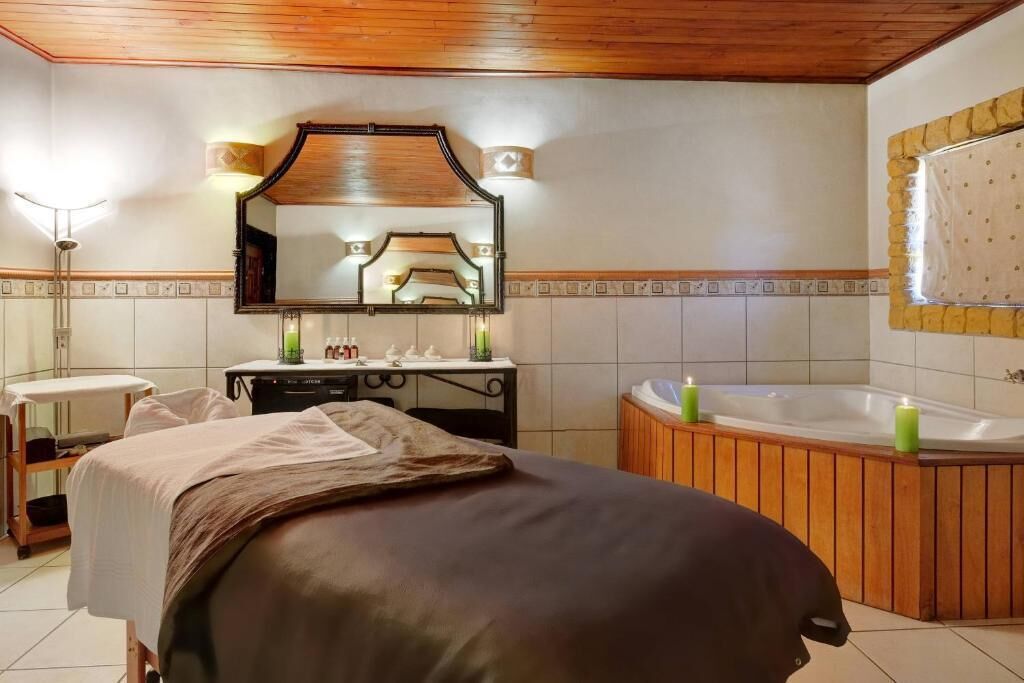 A hotel room with a bed, a wooden vanity, a large mirror, a bathtub, and a window with a curtain.