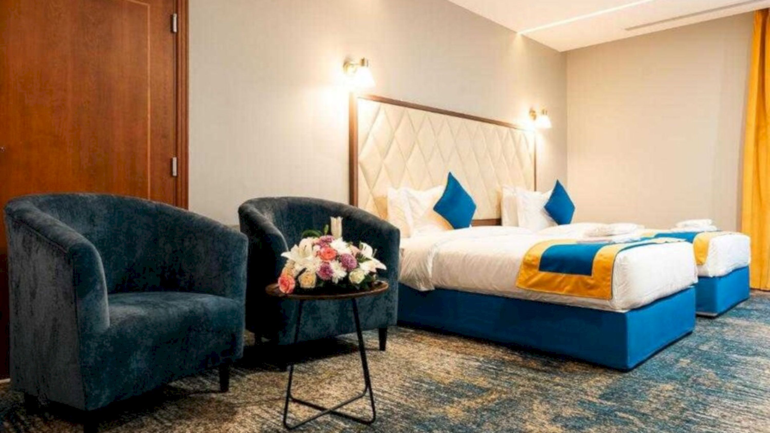 A hotel room with a bed, two armchairs, a small table with flowers, and a nightstand.