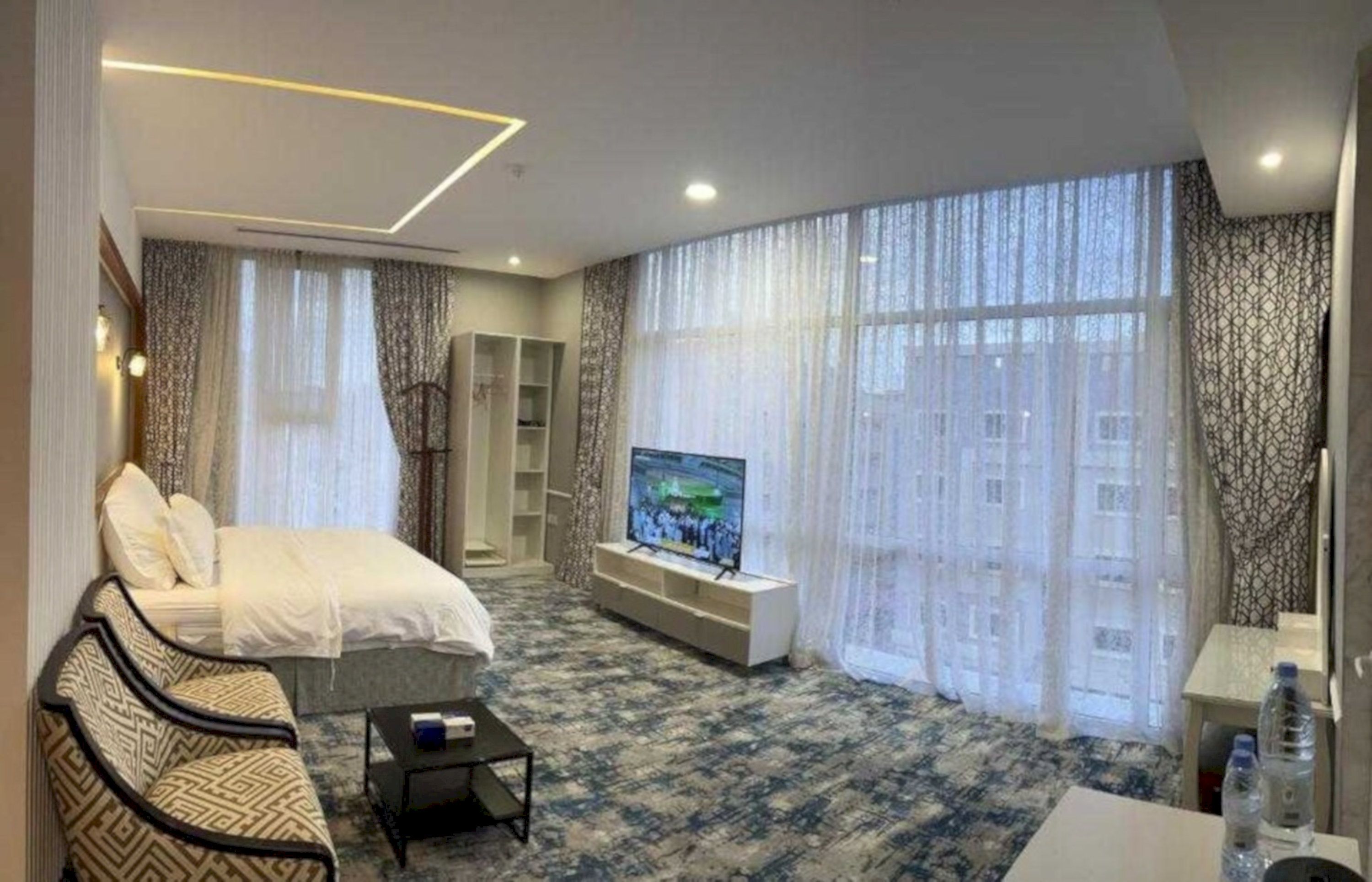 A hotel room with a bed, a TV, a sofa, a coffee table, and a large window with curtains.