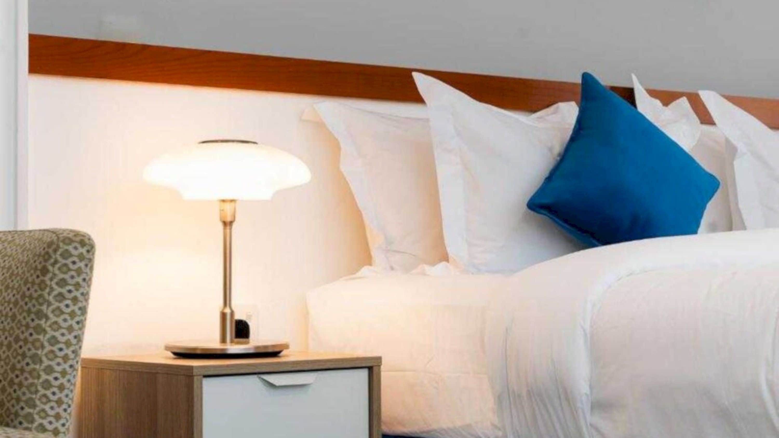 A hotel room with a bed, white bedding, a nightstand with a lamp, and a chair.