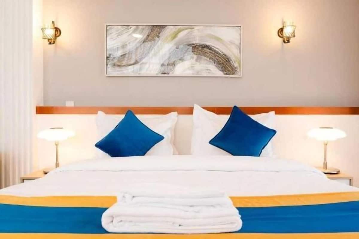 A hotel room with a bed, two wall-mounted lamps, a painting, and a folded towel on the bed.