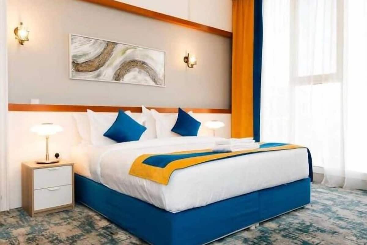 A hotel room with a bed, a nightstand, a wall-mounted artwork, and a window with curtains.