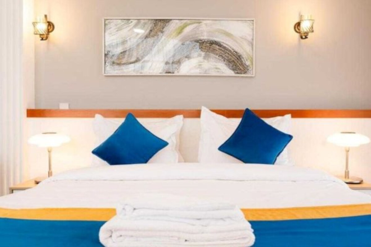 A hotel room with a bed, two bedside lamps, a painting on the wall, and a folded towel on the bed.