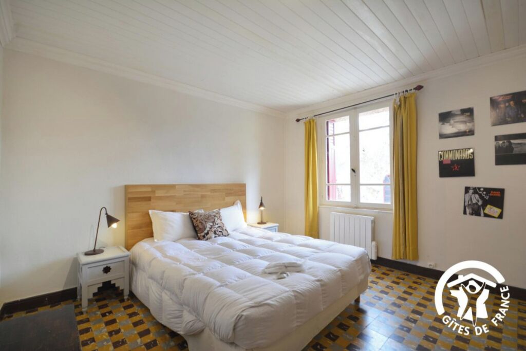 Een slaapkamer met een houten hoofdeinde, een bed met witte beddengoed, een nachtkastje met een lamp, een raam met gele gordijnen en een radiator.