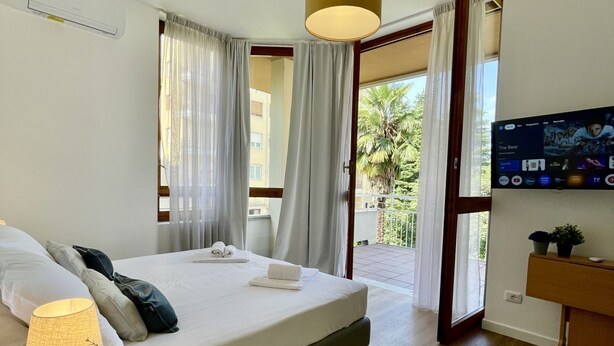 A modern hotel room with a bed, a TV, a desk, and a balcony with curtains.