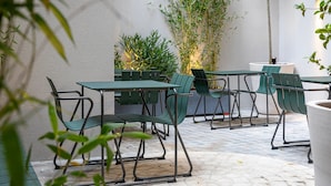 Een terras buiten met groene metalen stoelen en tafels, omgeven door potplanten en een met textuur bewerkte muur.