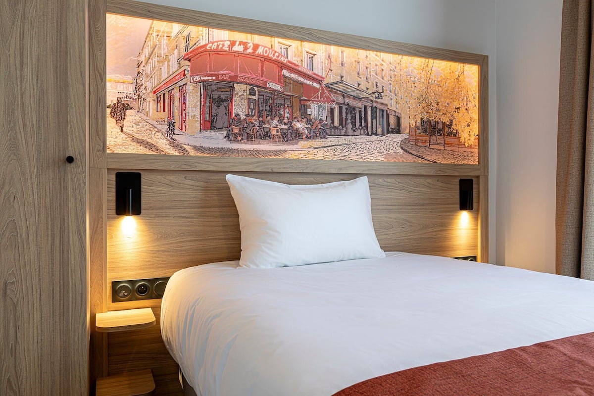 A hotel room with a bed, a white pillow, a headboard with a painting of a café, and two bedside lamps.